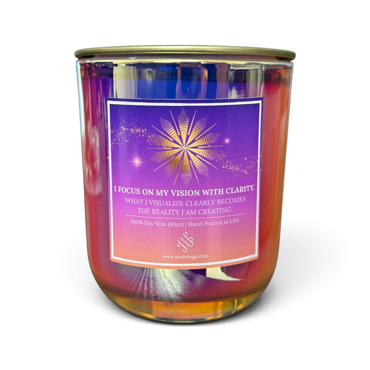 A 16 oz soy wax candle in a clear glass jar with a gold-toned metal lid, featuring a purple and pink label with a gold starburst design and the text 'I FOCUS ON MY VISION WITH CLARITY.