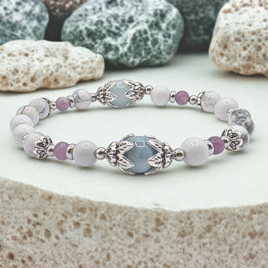 A beaded stretch bracelet featuring light blue, white, and lavender beads with silver-toned metal accents, displayed on a textured stone surface with decorative rocks in the background.