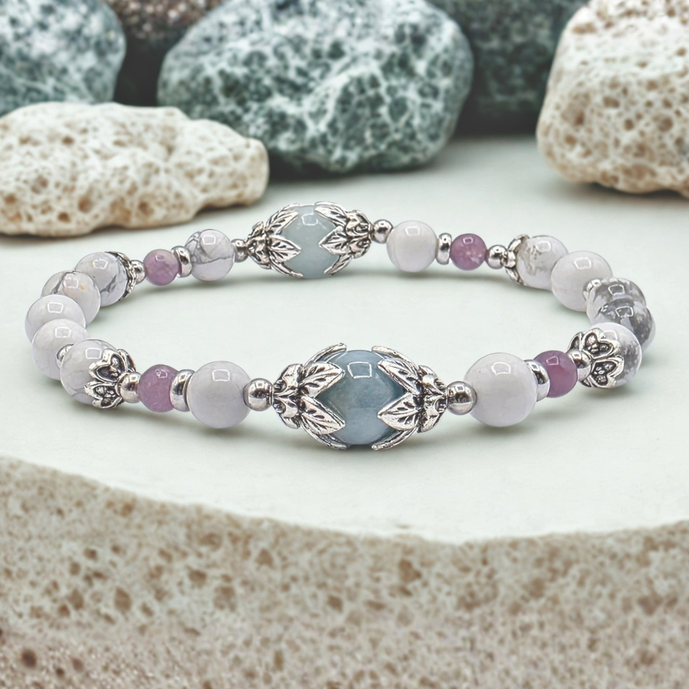A beaded stretch bracelet featuring light blue, white, and lavender beads with silver-toned metal accents, displayed on a textured stone surface with decorative rocks in the background.